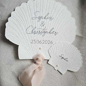 May include: White seashell-shaped wedding place cards with embossed details. The larger card has the names "Sophia & Christopher" and the date "25.06.2026" in black script. A smaller card reads "Sophie". A pink ribbon and pearl accents add a delicate touch.