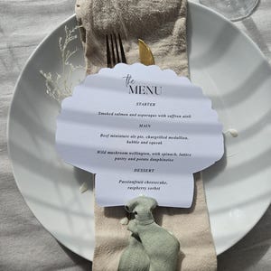 May include: A restaurant table setting with a white plate, beige napkin, and a menu card. The menu card displays the text "the MENU" and a list of dishes, including smoked salmon and passionfruit cheesecake. A gold knife and black fork are visible.