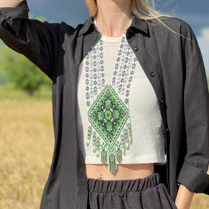 May include: A woman wearing a black shirt and a straw hat with a black band. She is wearing a white top with a green and white beaded necklace. The necklace has a diamond-shaped design with a green floral pattern.