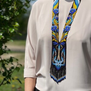 May include: A beaded necklace with a black base, featuring blue and yellow floral patterns. The necklace has a long, fringed pendant and is worn against a light-colored blouse. The design incorporates elements of the Ukrainian flag.