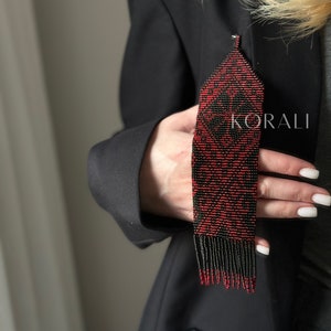 Könnte beinhalten: Ein roter und schwarzer Perlenfransenanhänger mit geometrischem Muster. Der Anhänger hängt an einer schwarzen Nadel und wird von einer Hand gehalten.