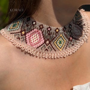Puede incluir: Un collar tipo gargantilla ancho con intrincados abalorios en tonos rosa pálido, amarillo, verde y plata metálica. El diseño incluye patrones geométricos de diamantes y hexágonos con un fleco de pequeñas cuentas rosa pálido en el borde.