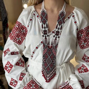 May include: A white embroidered blouse with red floral patterns on the sleeves and cuffs. A long beaded necklace with a geometric design in red, black, and white hangs over the blouse. The necklace has a fringe of red beads at the bottom. The word "KORALI" is visible.