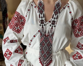 May include: A white embroidered blouse with red floral patterns on the sleeves and cuffs. A long beaded necklace with a geometric design in red, black, and white hangs over the blouse. The necklace has a fringe of red beads at the bottom. The word "KORALI" is visible.