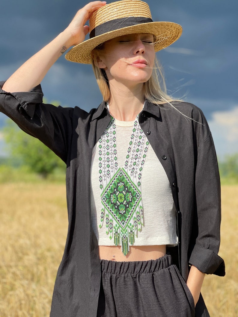 May include: A woman wearing a black shirt and a straw hat with a black band. She is wearing a white top with a green and white beaded necklace. The necklace has a diamond-shaped design with a green floral pattern.