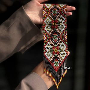 May include: A long, beaded necklace with a geometric pattern in red, white, black, green, and yellow. The necklace has a fringe of black beads with red and yellow accents.