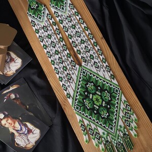 May include: A long, white and green beaded necklace with a geometric pattern and fringe. The necklace is displayed on a wooden surface.
