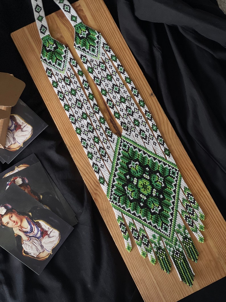 May include: A long, white and green beaded necklace with a geometric pattern and fringe. The necklace is displayed on a wooden surface.