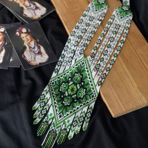 May include: A long, white and green beaded necklace with a geometric pattern. The necklace has a fringe at the bottom and is made of small, round beads.