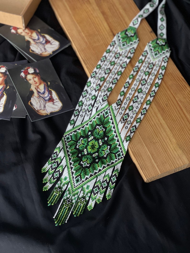 May include: A long, white and green beaded necklace with a geometric pattern. The necklace has a fringe at the bottom and is made of small, round beads.