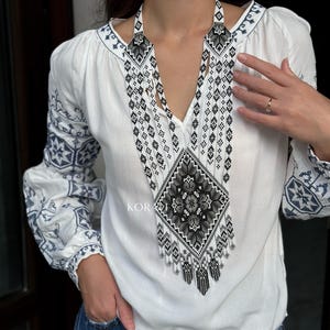 May include: A white embroidered blouse with a long, black and white beaded necklace. The necklace features a geometric pattern and a fringe.