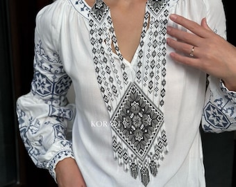 May include: A white embroidered blouse with a long, black and white beaded necklace. The necklace features a geometric pattern and a fringe.