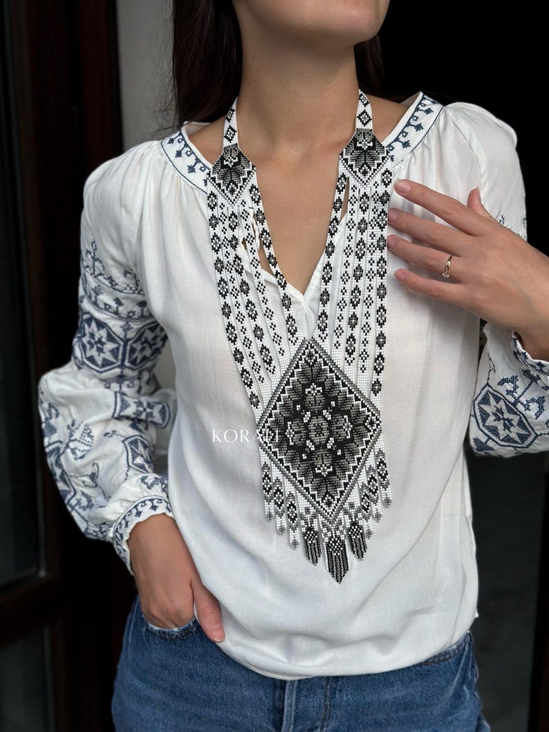 May include: A white embroidered blouse with a long, black and white beaded necklace. The necklace features a geometric pattern and a fringe.