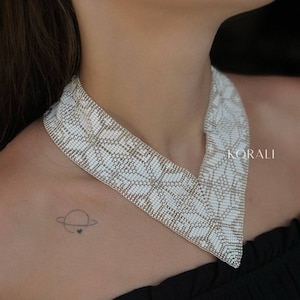 Może przedstawiać: Biały naszyjnik choker z koralików w kształcie litery V. Naszyjnik wykonany jest z małych, białych koralików i ma srebrne wykończenie. Naszyjnik nosi osoba z małym tatuażem planety na klatce piersiowej.