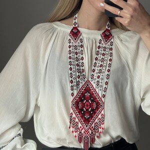 May include: A white beaded necklace with a red and black embroidered design. The necklace is long and has a fringe at the bottom.