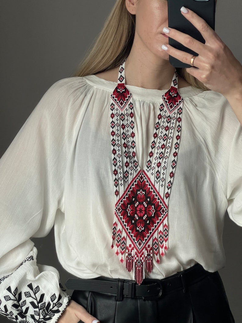 May include: A white beaded necklace with a red and black embroidered design. The necklace is long and has a fringe at the bottom.