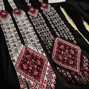 May include: Three long beaded necklaces with intricate red, white, and black geometric patterns. The necklaces have a fringe of beads at the bottom.