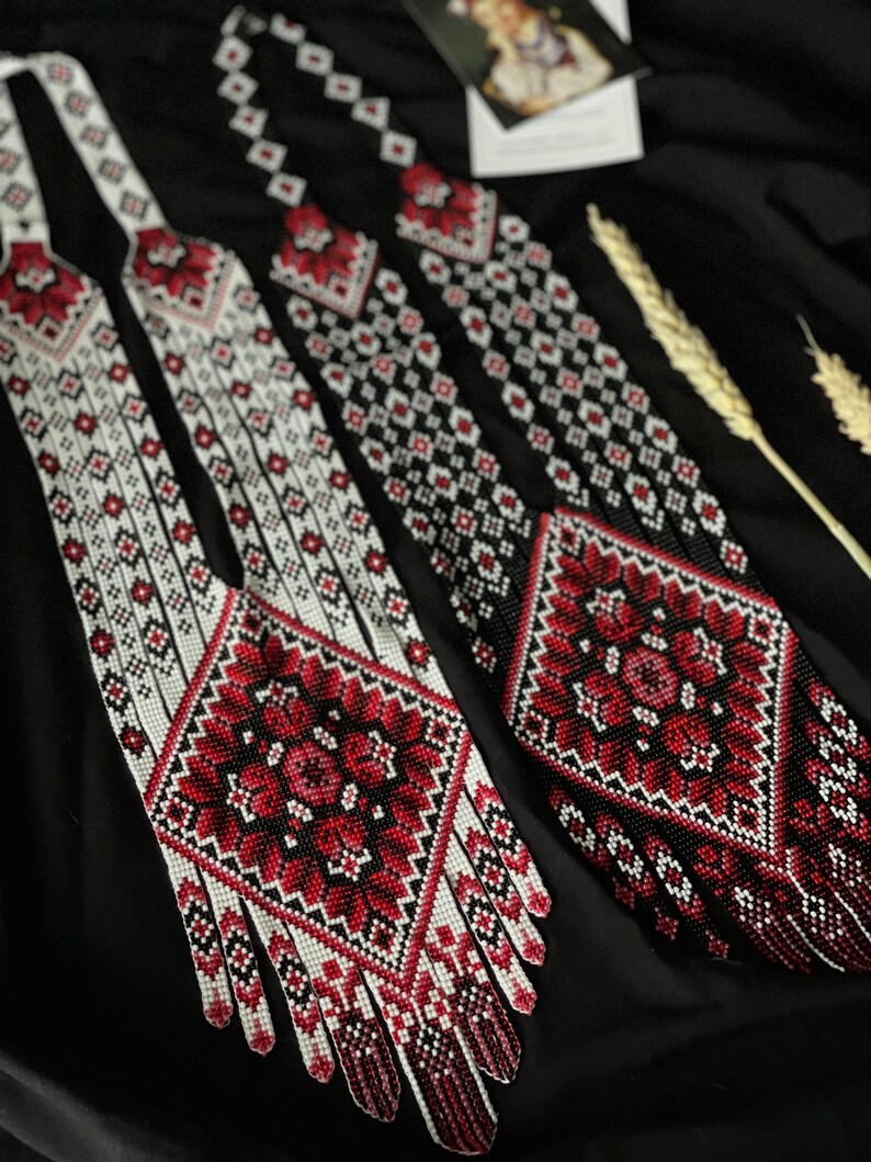 May include: Three long beaded necklaces with intricate red, white, and black geometric patterns. The necklaces have a fringe of beads at the bottom.