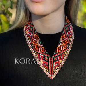 May include: A black v-neck shirt with a colourful beaded necklace. The necklace features geometric patterns in red, orange, yellow, green, white and black.