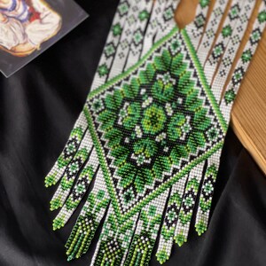 May include: A green and white beaded necklace with a geometric pattern and fringe. The necklace is made of small beads and has a traditional design.