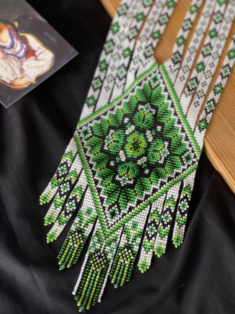 May include: A green and white beaded necklace with a geometric pattern and fringe. The necklace is made of small beads and has a traditional design.
