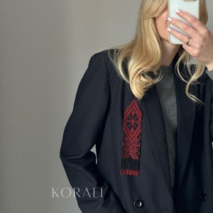 Op de afbeelding: Een zwart blazer met een rode en zwarte kralenhanger met een geometrisch patroon. De hanger hangt aan de halslijn van de blazer. De tekst "KORALI" is zichtbaar op de blazer.