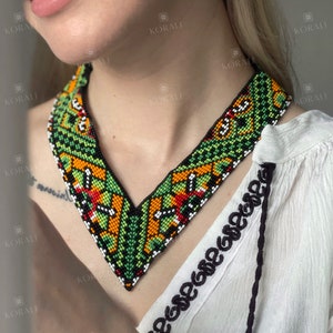 May include: A green, orange, black, and white beaded necklace with a V-shaped design. The necklace is worn by a person with a white shirt.