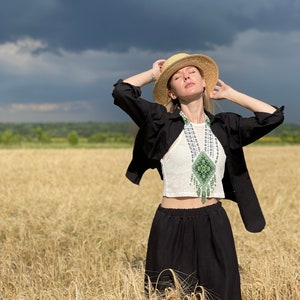 May include: A woman wearing a black shirt and black pants stands in a field of wheat. She is wearing a straw hat and a beaded necklace with green and white accents. The sky is cloudy.