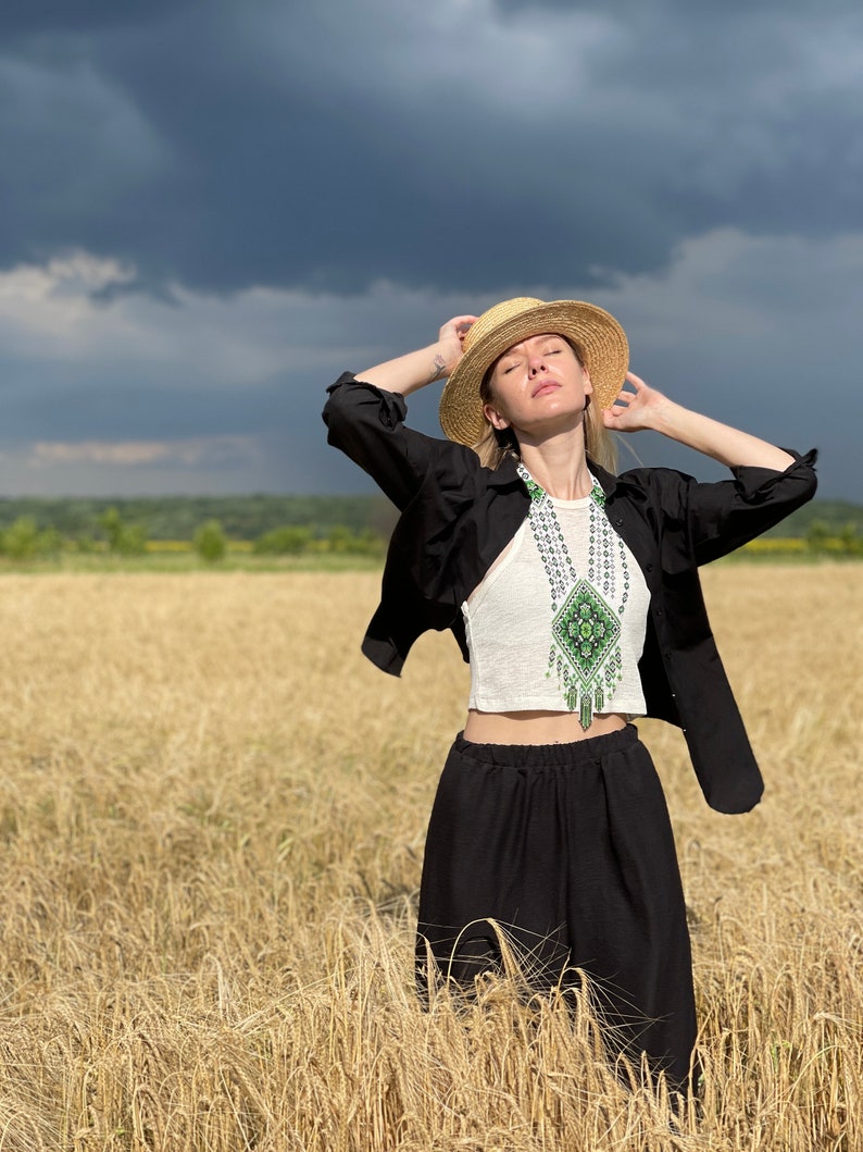 May include: A woman wearing a black shirt and black pants stands in a field of wheat. She is wearing a straw hat and a beaded necklace with green and white accents. The sky is cloudy.