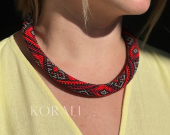 May include: A red, black, and white beaded necklace with a geometric pattern. The necklace is worn by a person with a light yellow shirt.