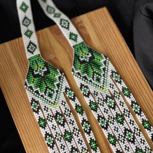 May include: A white and green beaded necklace with a geometric pattern. The necklace is made of small beads and has a fringe at the bottom.