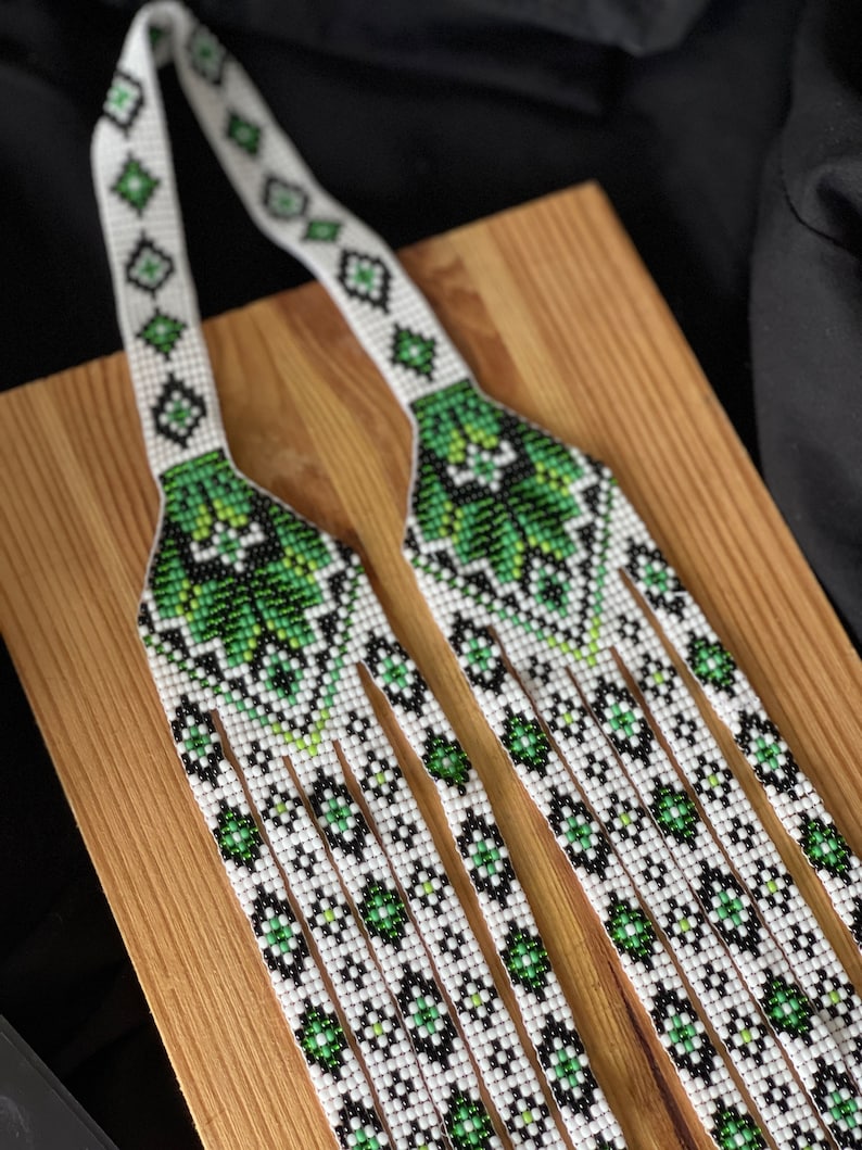 May include: A white and green beaded necklace with a geometric pattern. The necklace is made of small beads and has a fringe at the bottom.