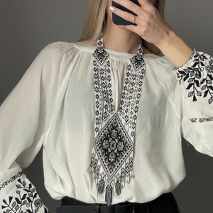 May include: A white blouse with black embroidered floral designs on the sleeves. The blouse is worn with a black leather belt and a long, black and white beaded necklace with a geometric pattern.