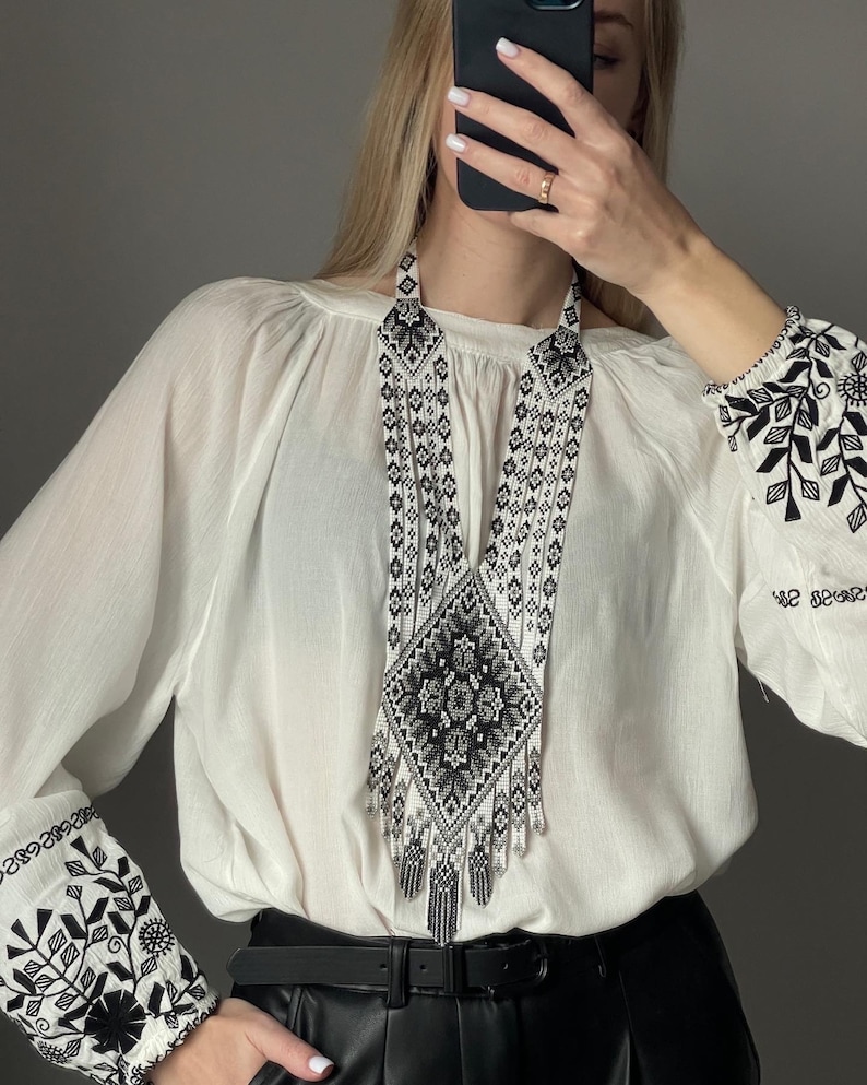 May include: A white blouse with black embroidered floral designs on the sleeves. The blouse is worn with a black leather belt and a long, black and white beaded necklace with a geometric pattern.