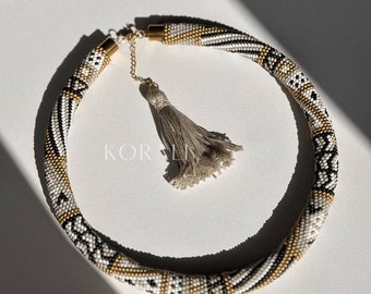 May include: A white, black, and gold beaded necklace with a geometric pattern. The necklace has a tassel on the end.