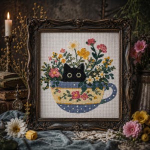 以下が含まれることがあります： 暗い装飾フレームに入ったクロスステッチ刺繍。黄色と青の花柄のティーカップから顔を出す黒猫が描かれており、カップの中にはピンク、黄色、白の花々が活けられています。背景は明るい色の布地です。