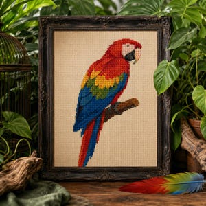 May include: A cross-stitch embroidery of a scarlet macaw perched on a branch, displayed in a dark ornate wooden frame. The bird features vibrant red, yellow, and blue plumage against a neutral beige fabric background with visible grid texture.