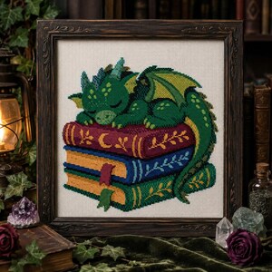 Puede incluir: Un bordado de punto de cruz que muestra un pequeño dragón verde durmiendo sobre una pila de tres libros. Los libros son de color burdeos, azul y amarillo, decorados con patrones de hojas doradas y marcapáginas. La obra está en un marco de madera oscura.