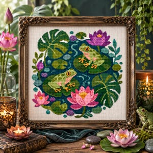 May include: A framed cross-stitch embroidery featuring two light green frogs resting on lily pads within a dark blue pond. The design includes vibrant pink and purple lotus flowers, large green monstera leaves, and small circular water ripples.