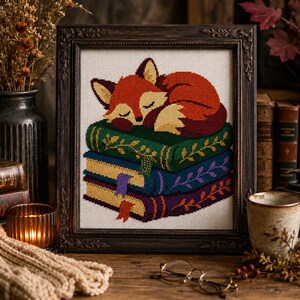 May include: A framed cross-stitch embroidery featuring a sleeping orange fox curled up on a stack of three books. The books are green, blue, and purple, each decorated with gold leaf patterns and small bookmarks. The frame is dark brown wood.