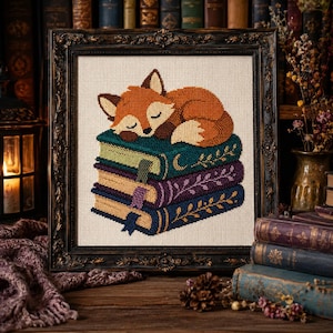 May include: A cross-stitch embroidery in a dark ornate frame featuring a sleeping orange fox resting on a stack of three books. The books are teal, purple, and blue, each decorated with gold leaf patterns and bookmarks, set against a cream fabric.