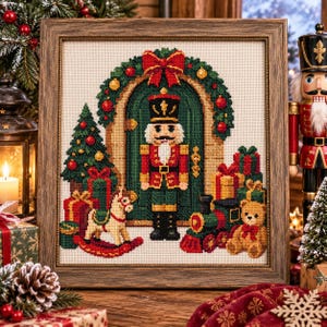 May include: A framed cross-stitch embroidery depicting a nutcracker standing before a green arched door adorned with a wreath. The scene includes a Christmas tree, a rocking horse, a toy train, a teddy bear, and wrapped gifts on a white fabric.