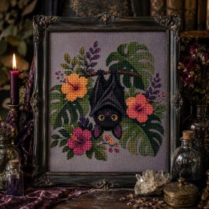 May include: A framed cross-stitch embroidery featuring a black bat hanging upside down, surrounded by vibrant orange, pink, and yellow hibiscus flowers, purple sprigs, and large green monstera leaves on a solid gray fabric background.