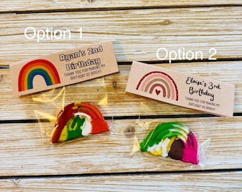 May include: Two clear plastic favor bags containing rainbow-shaped crayons. Option 1 features a kraft paper topper with a colorful rainbow and text reading Ryan's 2nd Birthday. Option 2 features a kraft paper topper with a muted rainbow and text reading Eloise's 3rd Birthday. Both include the phrase Thank you for making my birthday so bright.