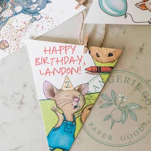 May include: A set of triangular paper bunting flags featuring watercolor illustrations of a gray cat with confetti, a pig with balloons, and a mouse wearing a party hat with the text HAPPY BIRTHDAY, LANDON! The flags are attached to twine with small wooden clips.