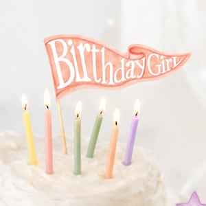 May include: A white frosted cake with lit candles and a "Birthday Girl" banner. Cupcakes with star and rainbow toppers are arranged around the cake. The candles are pastel colors.