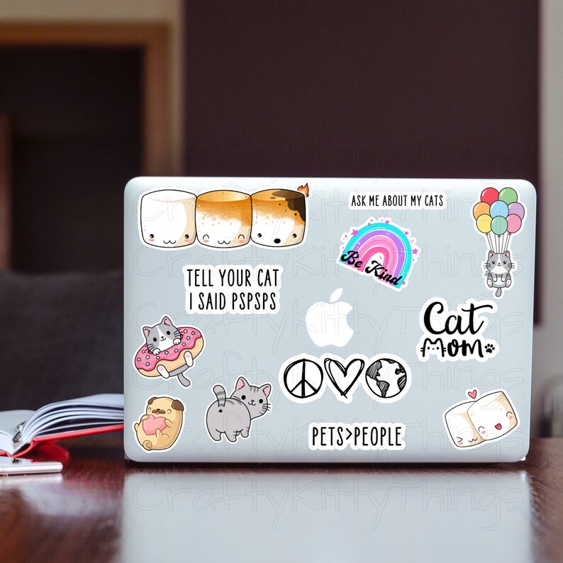 May include: A laptop computer with various stickers, including a cat mom sticker, a peace sign, a heart, a globe, a rainbow with the text "Be Kind", a cat in a donut, a cat with the text "Tell your cat I said pspsps", three marshmallows, a cat with a hot air balloon, and the text "Ask me about my cats".