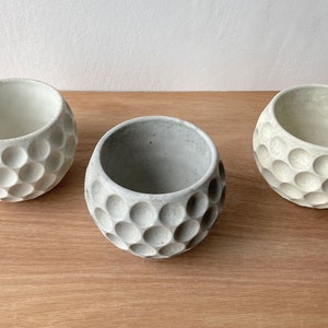 May include: Three white ceramic planters with a textured, dimpled surface, resembling a golf ball. The planters are arranged in a row on a wooden surface.