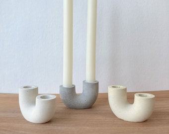 May include: Three concrete candle holders in the shape of a U. The holders are in different shades of gray and white. Two of the holders are empty, and the third holder has two white candles.