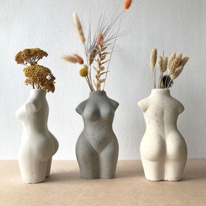 May include: Three concrete vases in the shape of a female torso, each filled with dried flowers. The vases are grey and off-white. The dried flowers include yellow, orange, and beige tones, providing a natural contrast to the concrete.